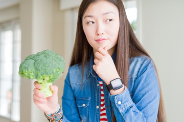 Beautiful Asian woman eating green fresh broccoli serious face thinking about question, very...