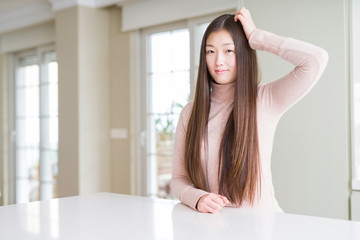 Beautiful Asian woman wearing casual sweater on white table confuse and wonder about question. Uncertain with doubt, thinking with hand on head. Pensive concept.