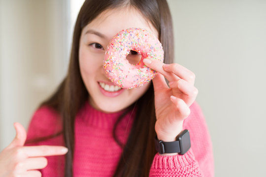 Beautiful Asian woman eating pink sugar donut with surprise face pointing finger to himself