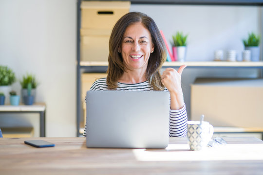 Middle age senior woman sitting at the table at home working using computer laptop smiling with happy face looking and pointing to the side with thumb up.