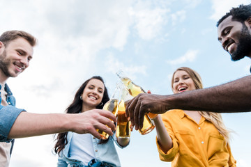 low angle view of cheerful multicultural men and women clinking bottles with beer