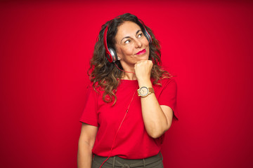 Middle age senior woman wearing headphones listening to music over red isolated background with hand on chin thinking about question, pensive expression. Smiling with thoughtful face. Doubt concept.