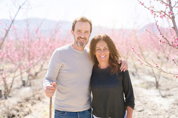 Beautiful middle age senior couple smiling in love at romantic garden of peach trees with pink petals on a sunny day of spring