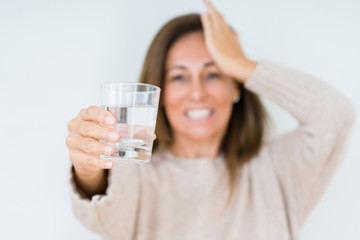 Middle age woman drinking glass of water isolated background stressed with hand on head, shocked with shame and surprise face, angry and frustrated. Fear and upset for mistake.