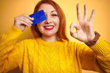 Young beautiful redhead woman holding credit card over yellow isolated background doing ok sign with fingers, excellent symbol