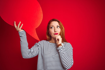 Young beautiful redhead woman holding speech bubble over red isolated background serious face...