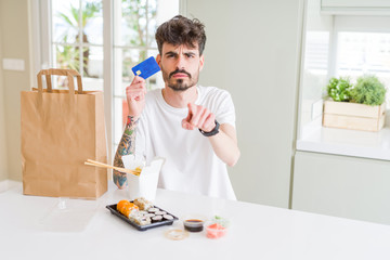 Young man eating asian sushi from home delivery using credit card as payment pointing with finger to the camera and to you, hand sign, positive and confident gesture from the front