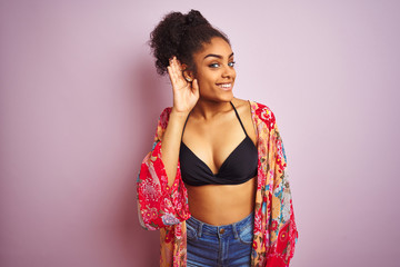 American woman on holiday wearing bikini and colorful dress over isolated pink background smiling with hand over ear listening an hearing to rumor or gossip. Deafness concept.