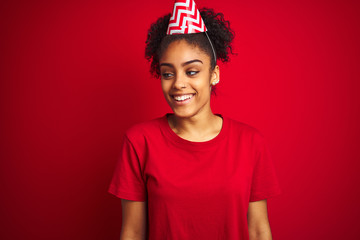 Young afro american woman wearing birthday hat over isolated red background looking away to side with smile on face, natural expression. Laughing confident.