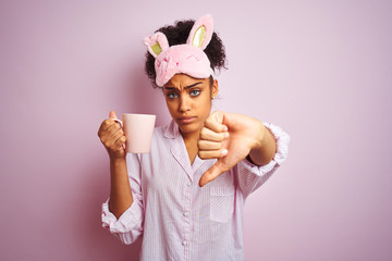 Afro woman wearing pajama and mask drinking a cup of coffee over isolated pink background with...