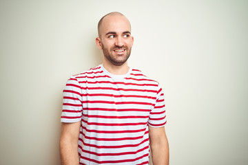 Young bald man with beard wearing casual striped red t-shirt over white isolated background looking away to side with smile on face, natural expression. Laughing confident.