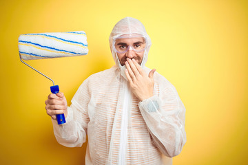 Young man wearing painter equipment and holding painting roller over isolated yellow background cover mouth with hand shocked with shame for mistake, expression of fear, scared in silence