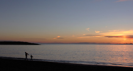 Fototapeta premium Sunset at ocean with silhouette of man and boy walking along beach with copy space