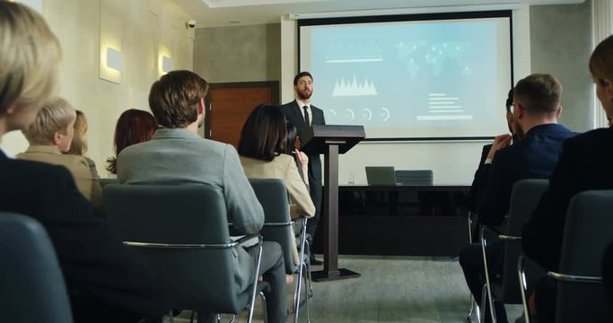 Young Caucasian male worker or head of department standing at the lecturn and having a report with demonstration charts on the screen behind him.