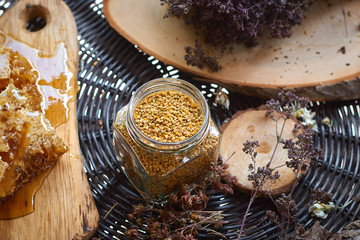 A table with a wicker table top, honey in honeycombs on a wooden board, honey in an open glass jar, with medicinal herbs oregano, chamomile and strawberry flowers. flower pollen collected by bees