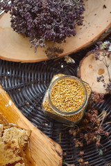 A table with a wicker table top, honey in honeycombs on a wooden board, honey in an open glass jar, with medicinal herbs oregano, chamomile and strawberry flowers. flower pollen collected by bees