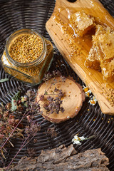 A table with a wicker table top, honey in honeycombs on a wooden board, honey in an open glass jar, with medicinal herbs oregano, chamomile and strawberry flowers. flower pollen collected by bees
