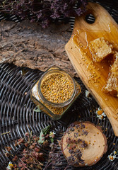 A table with a wicker table top, honey in honeycombs on a wooden board, honey in an open glass jar, with medicinal herbs oregano, chamomile and strawberry flowers. flower pollen collected by bees