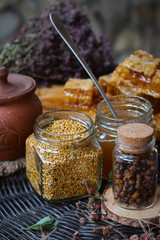 A table with a wicker table top, honey in honeycombs on a wooden board, honey in an open glass jar, with medicinal herbs oregano, chamomile and strawberry flowers. flower pollen collected by bees