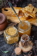 A table with a wicker table top, honey in honeycombs on a wooden board, honey in an open glass jar, with medicinal herbs oregano, chamomile and strawberry flowers. flower pollen collected by bees