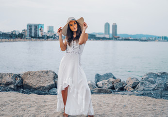 Pretty woman wearing white dress and pamela hat standing over the city beach, sea. Happy summer...