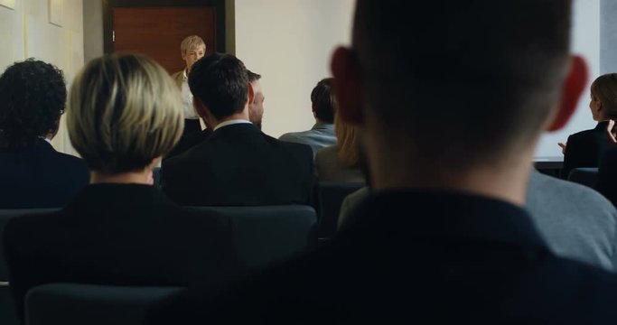 View over the heads of the audience on the Caucasian woman business coach opening a door and entering the aula, then going to the lecturn and starting her speech.