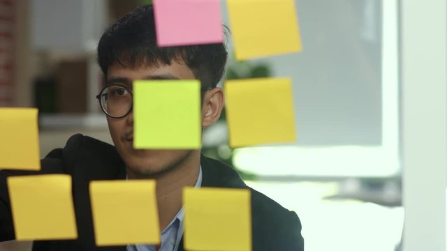 Asian creative man stick a sticky note on glass board. Young professional business male write information, strategy, reminder on paper, business situation, startup in Loft office concept. Slow motion.