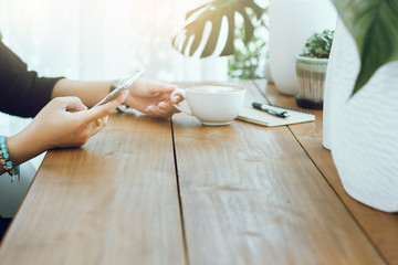 Closeup of girl hands using smartphone and holding cup of coffee with notebook on wooden table. selective focus soft and blurred background