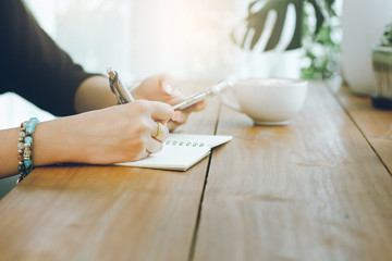 Closeup of hand young asian woman writing on notebook and use smartphone working with cup of coffee...