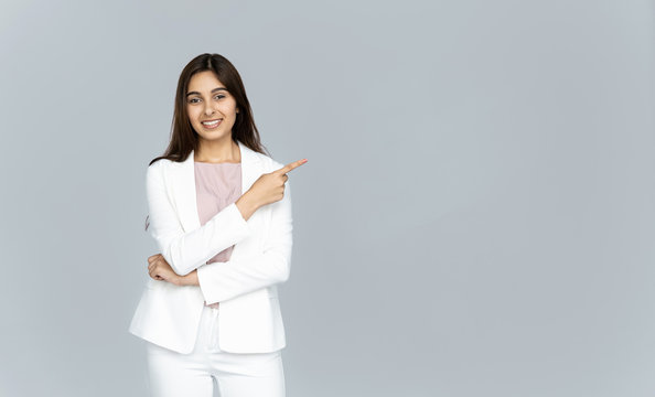 Happy indian young business woman wear white suit looking at camera pointing finger at copy space isolated on grey studio background, smiling beautiful confident hindu lady professional showing aside