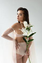 tender young woman in beige lingerie and mesh sleeves holding lilies behind back isolated on white