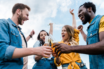 low angle view of happy multicultural men and women clinking bottles with beer