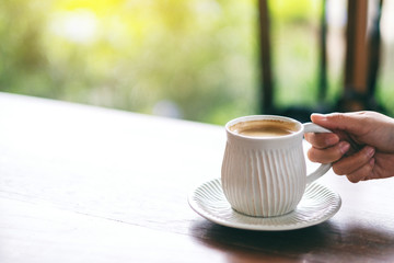 Closeup image of a hand holding a white cup of hot coffee on wooden table