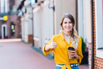 happy woman holding paper cup while standing on street and showing thumb up
