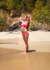 Fototapeta premium Young sporty woman in pink bikini swimwear and sunglasses posing on the wet sand beach looking to sun, hands in her hair, jungle bushes behind