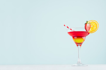 Summer fresh striped fruit cocktail with red and yellow liquor, orange slice, strawberry, straw in pastel mint color interior on white wood board.
