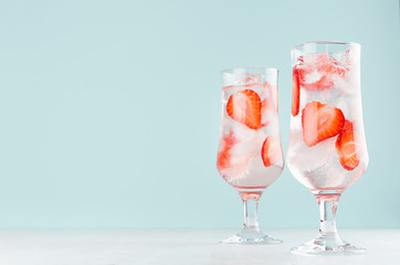 Fresh summer strawberry drink with red fruit pieces, ice cubes, tonic in two glasses light soft pastel  blue color background on white wood board.