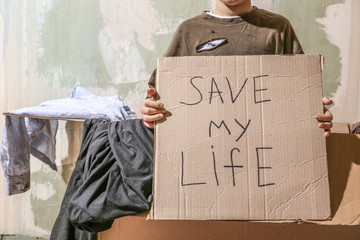 Homeless little boy holding piece of cardboard with text SAVE MY LIFE indoors