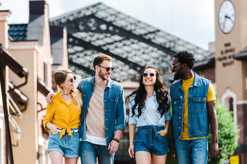 Obraz premium cheerful women with hands in pockets walking near multicultural men