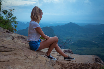 Woman taveler sitting on the cliff and looking at the beautiful landscape with mountains and valley with cloudy morning sky