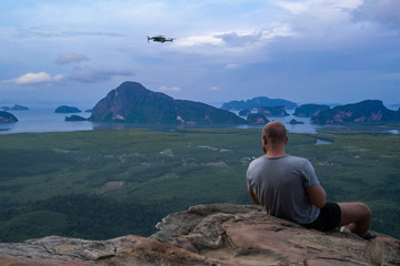 Back view of man drone operator sitting on the edge of the rock over beautiful green mountains and sky background