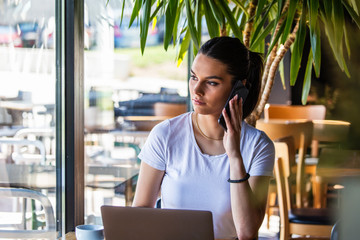 Young woman with coffee cup and laptop talking on her smart phone. Woman taking a break. Doing Business From the coffee shop