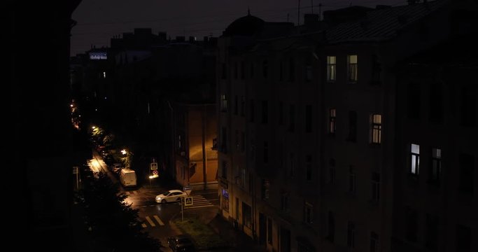 Top View Of Small Residential Street In A European Town At Nighttime. There Are Pedestrians And Cars Moving. Rainy Weather, Lightning On The Horizon Above The Roofs