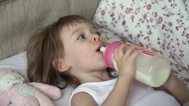 Portrait of little cute baby girl lies on bed waking up in morning takes bottle of milk, sucks and drinks with big appetite. Baby food formula concept