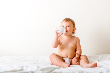Baby toddler sits on the white bed, smiles and drinks water from plastic bottle. Copy space