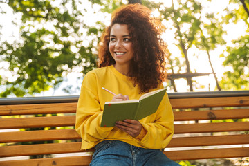 Happy young beautiful curly student girl sitting outdoors in nature park reading book writing notes.