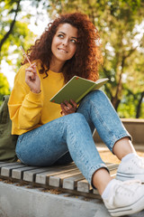Young beautiful curly student girl sitting outdoors in nature park writing notes in notebook.