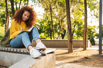 Young beautiful curly student girl sitting outdoors in nature park writing notes in notebook.