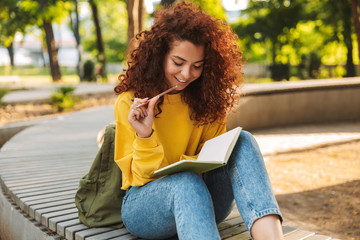 Young beautiful curly student girl sitting outdoors in nature park writing notes in notebook.