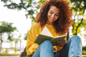 Young beautiful curly student girl sitting outdoors in nature park writing notes in notebook.